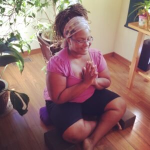 Dianne sits on a meditation cushion with hands in anjali mudra.